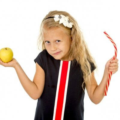 Motivation and autism: little beautiful female child with blond hair choosing dessert holding unhealthy but tasty red candy licorice and apple fruit in healthy versus unhealthy die nutrition isolated on white background